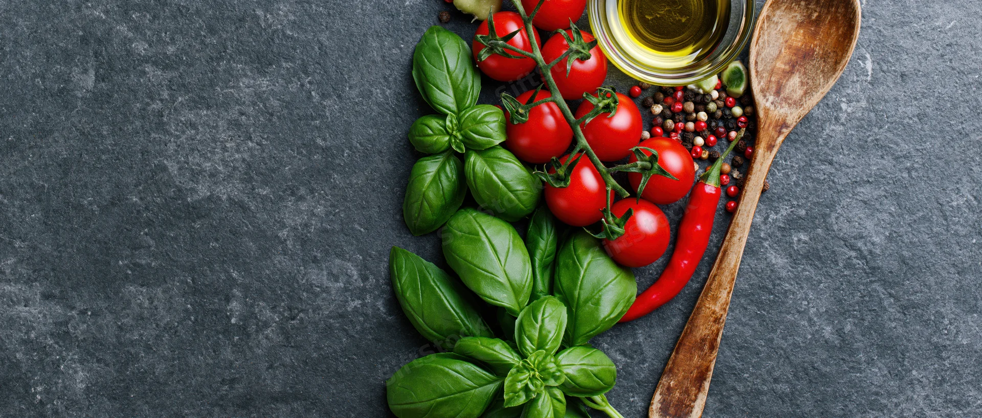 Fresh basil and cherry tomatoes on the vine with spices and olive oil arranged on dark slate surface