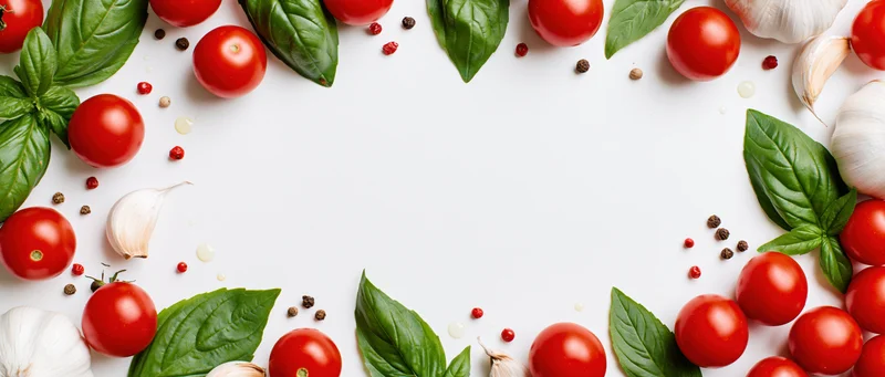 Cherry tomatoes, garlic bulbs and basil leaves arranged in a border around a white surface with copyspace
