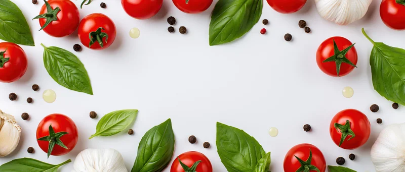 Cherry tomatoes, basil leaves, garlic bulbs, and peppercorns arranged in a border on white surface with central copyspace