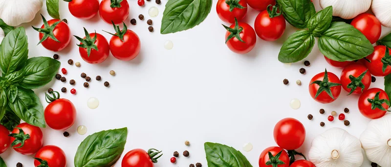 Cherry tomatoes, garlic bulbs and basil leaves arranged in a border around a white surface with copyspace