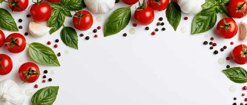 Cherry tomatoes, garlic bulbs, basil leaves, and peppercorns arranged in a border around a white surface with copyspace in the center