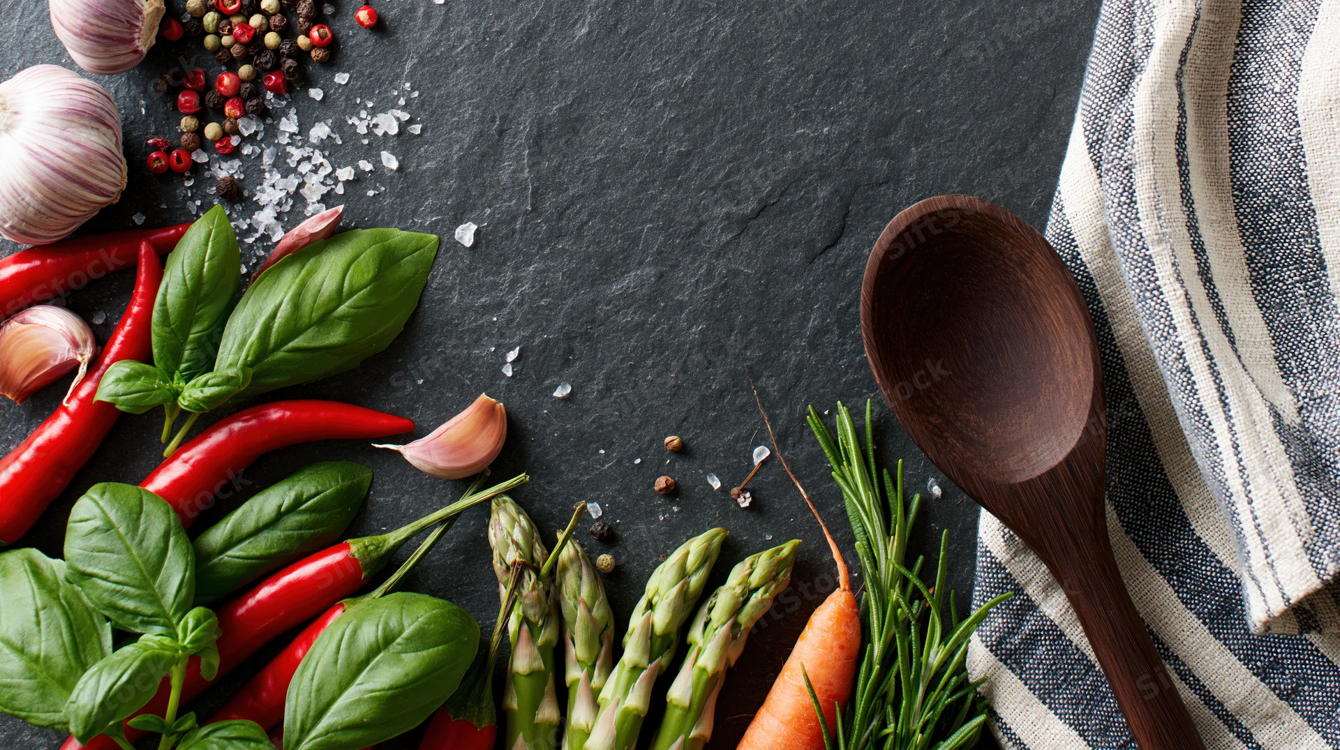 Fresh vegetables and herbs arranged with a wooden spoon on a dark slate surface