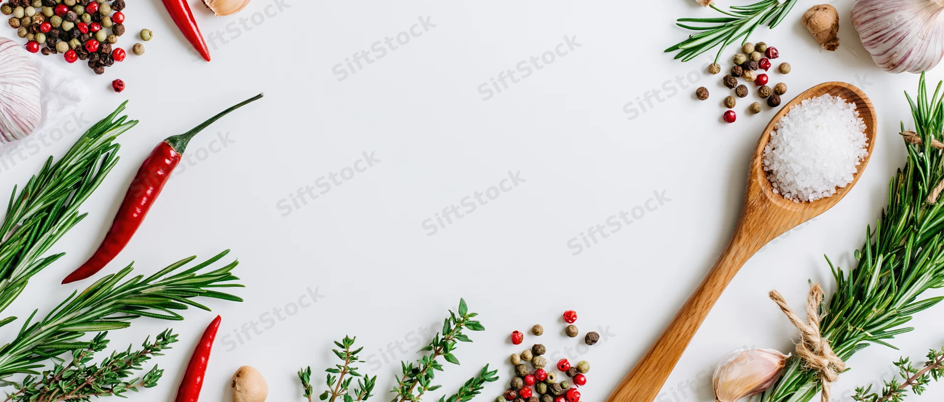 Fresh herbs and spices arranged around edges of white surface with wooden spoon holding salt