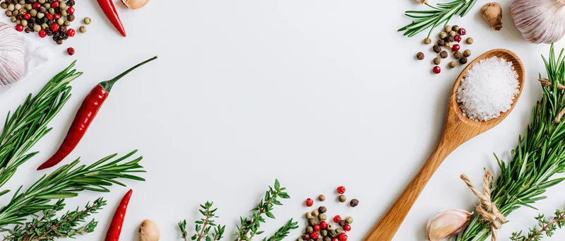 Fresh herbs and spices arranged around edges of white surface with wooden spoon holding salt