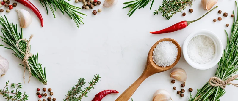Fresh herbs and spices arranged around edges of white marble surface with wooden spoon and bowl of salt in center