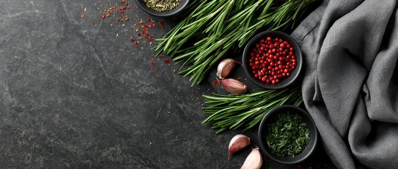 Fresh rosemary sprigs and small dark bowls of red peppercorns and dried herbs arranged on dark stone surface with gray cloth and garlic cloves