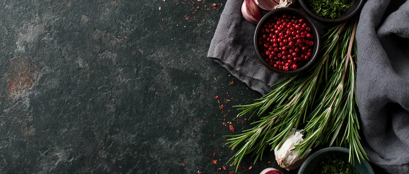 Fresh rosemary sprigs and bowls of red peppercorns and garlic arranged on dark textured surface with gray cloth and copyspace