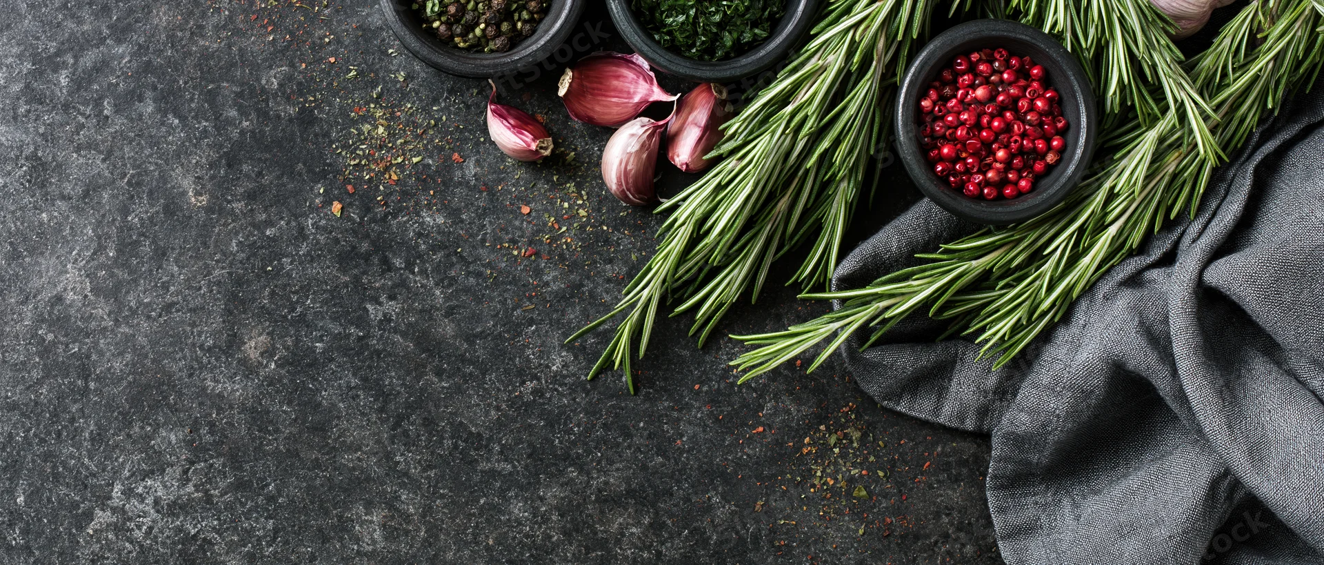 Fresh rosemary sprigs and small dark bowls of peppercorns and garlic arranged on a dark stone surface with a gray cloth, top view
