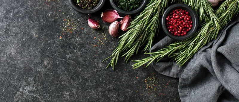 Fresh rosemary sprigs and small dark bowls of peppercorns and garlic arranged on a dark stone surface with a gray cloth, top view