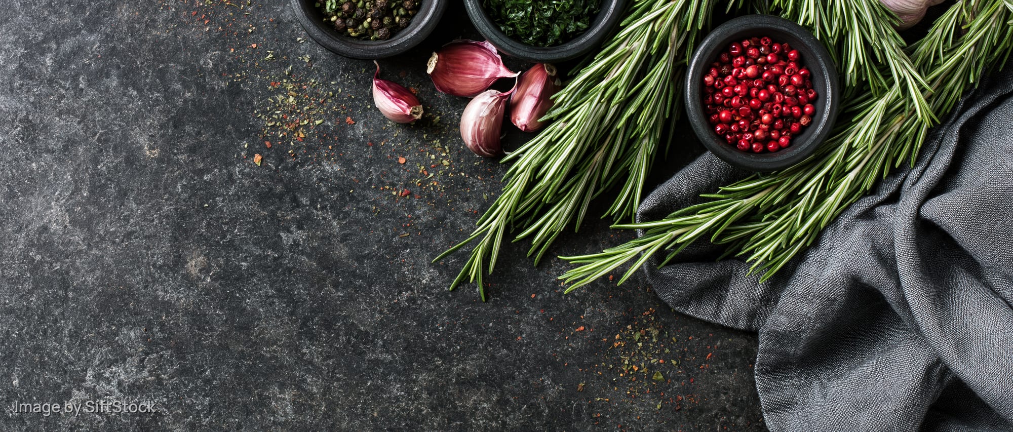 Fresh rosemary sprigs and small dark bowls of peppercorns and garlic arranged on a dark stone surface with a gray cloth, top view