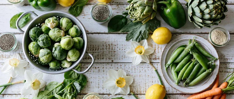 Fresh spring vegetables arranged in a flat lay on a whitewashed wooden table with daffodils and herbs