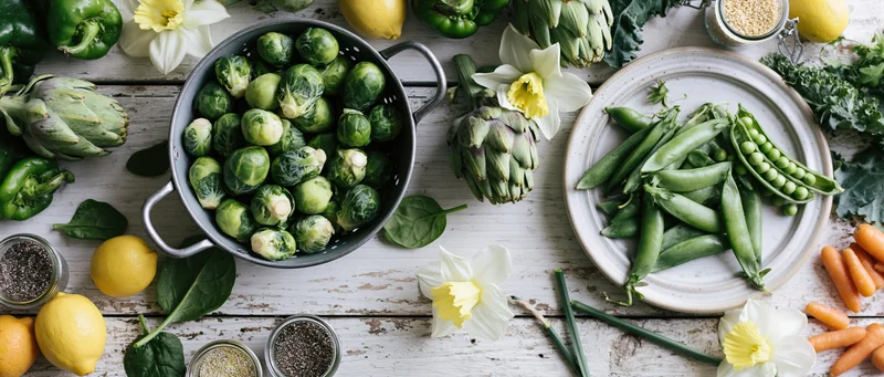 Fresh spring vegetables arranged in bowls and plates on a whitewashed wooden table with daffodils and lemons