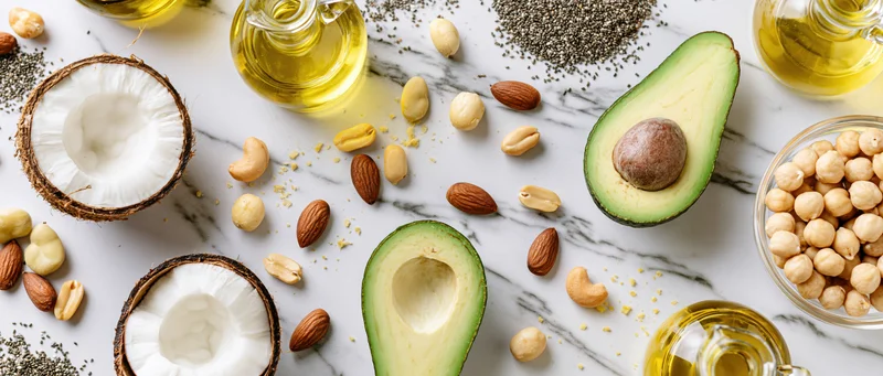 Healthy fats ingredients including avocado, coconut, nuts and seeds arranged on white marble surface in flat lay composition