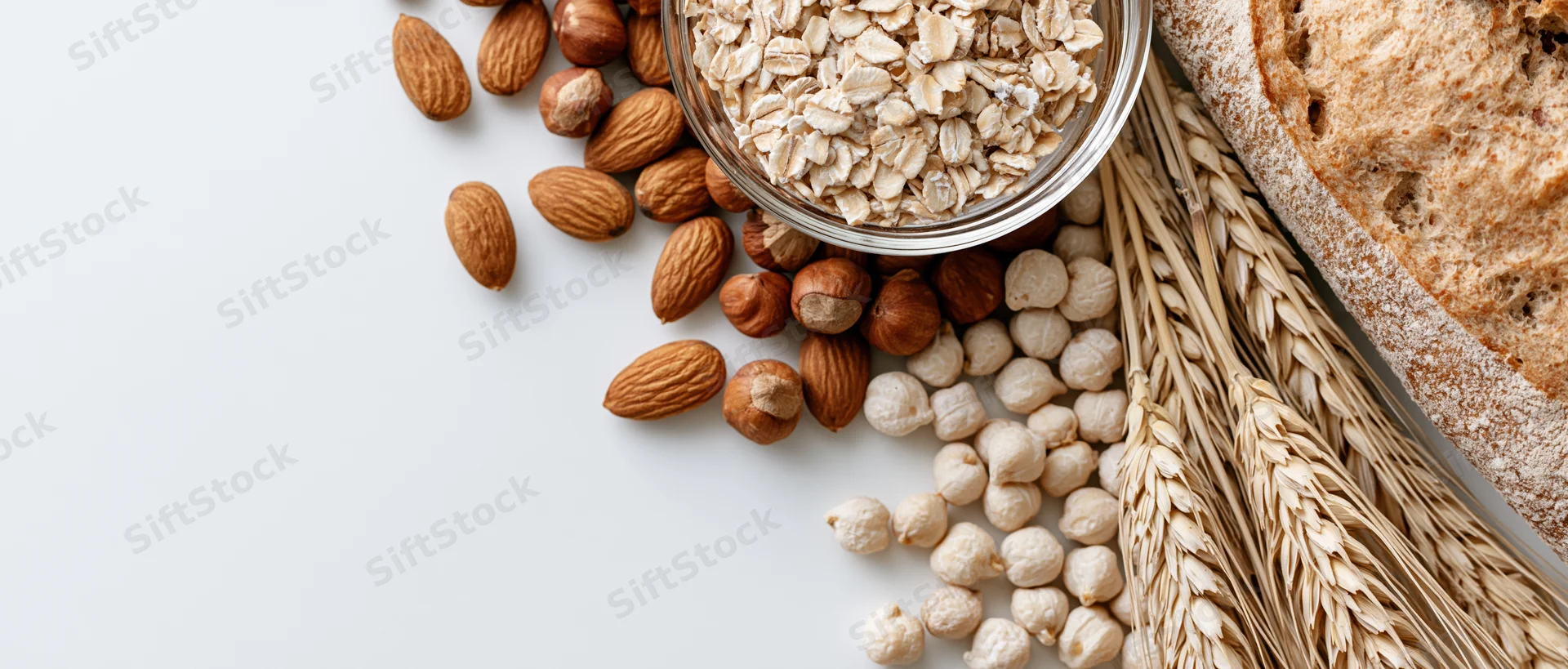 Flat lay of oats, nuts, chickpeas, wheat stalks and bread on white surface