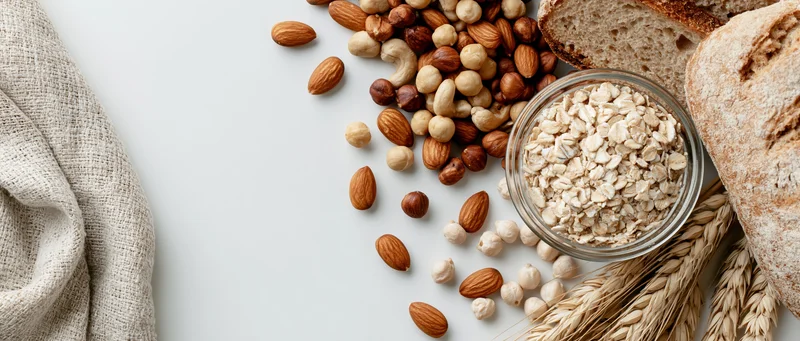 Mixed nuts and rolled oats in a glass bowl with bread and wheat stalks arranged on a light surface