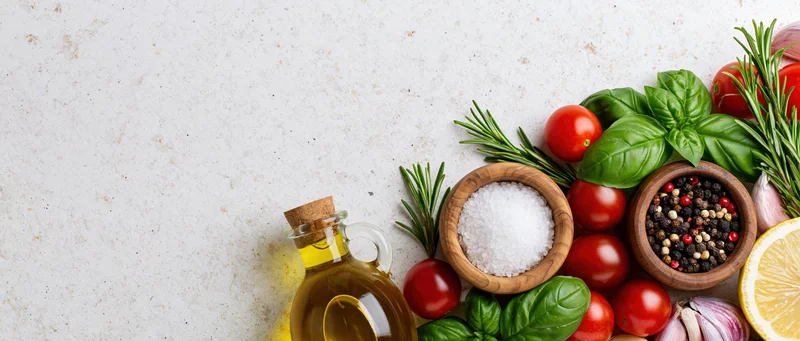 Fresh vegetables herbs and spices arranged with olive oil and salt on a light countertop