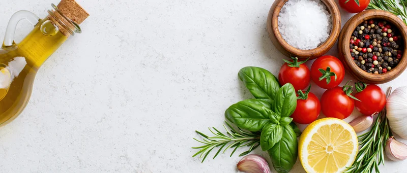 Fresh ingredients including tomatoes, basil, lemon, garlic, rosemary, salt and pepper arranged on a white countertop with olive oil bottle on the side