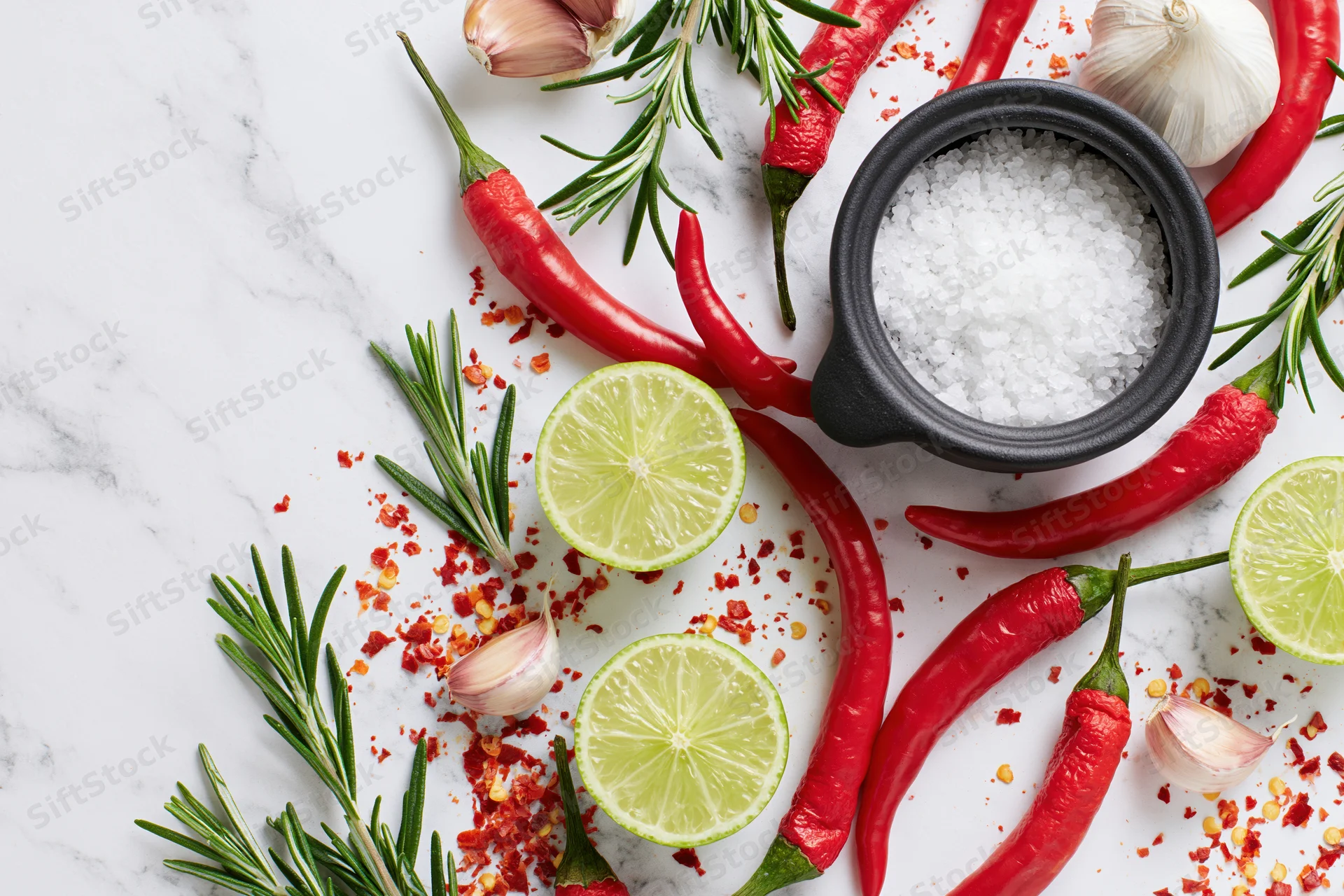 Red chili peppers, halved limes, garlic cloves, rosemary, and salt arranged on a marble surface in a flat lay composition