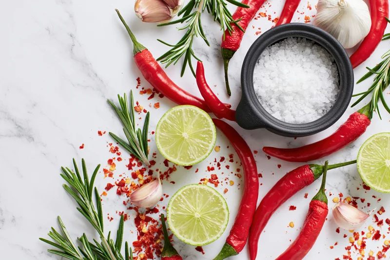 Red chili peppers, halved limes, garlic cloves, rosemary, and salt arranged on a marble surface in a flat lay composition