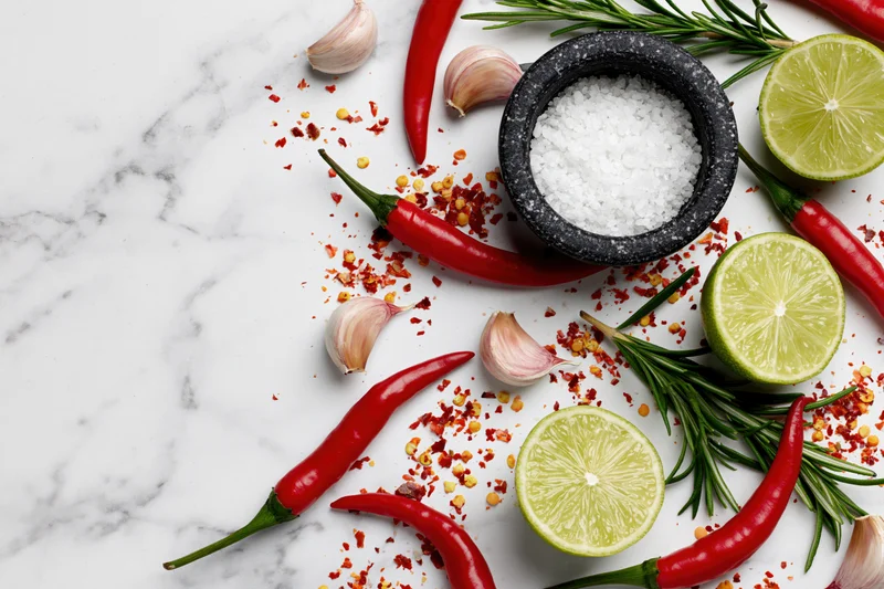 Red chili peppers, halved limes, garlic cloves, rosemary, and salt in a mortar arranged on a white marble surface
