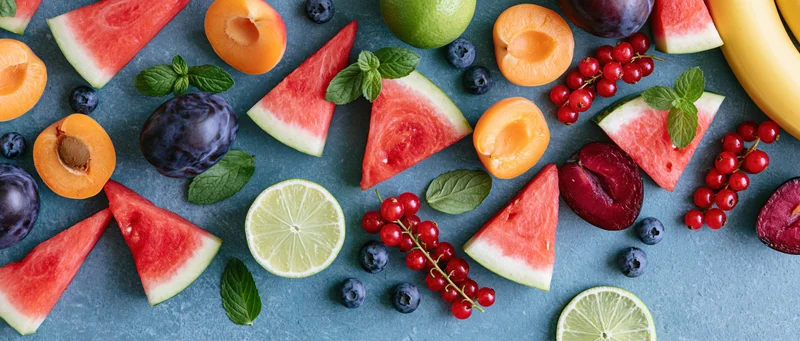Sliced watermelon wedges, halved apricots, plums, limes, bananas, redcurrants, blueberries, and mint leaves arranged on a gray slate surface in a flat lay top view