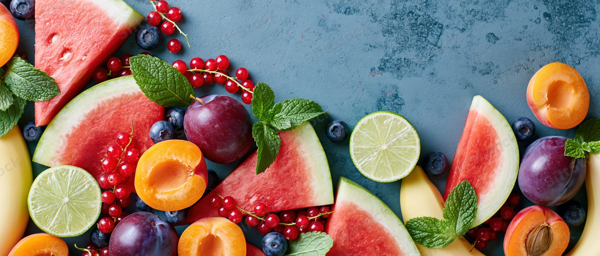Sliced watermelon wedges, halved apricots, plums, limes, and mint arranged on a dark textured surface with copyspace
