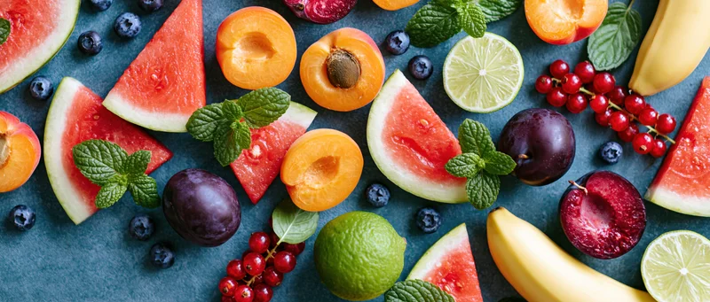 Sliced watermelon, apricots, plums, bananas, limes, blueberries, and mint arranged on dark slate surface in flat lay overhead view