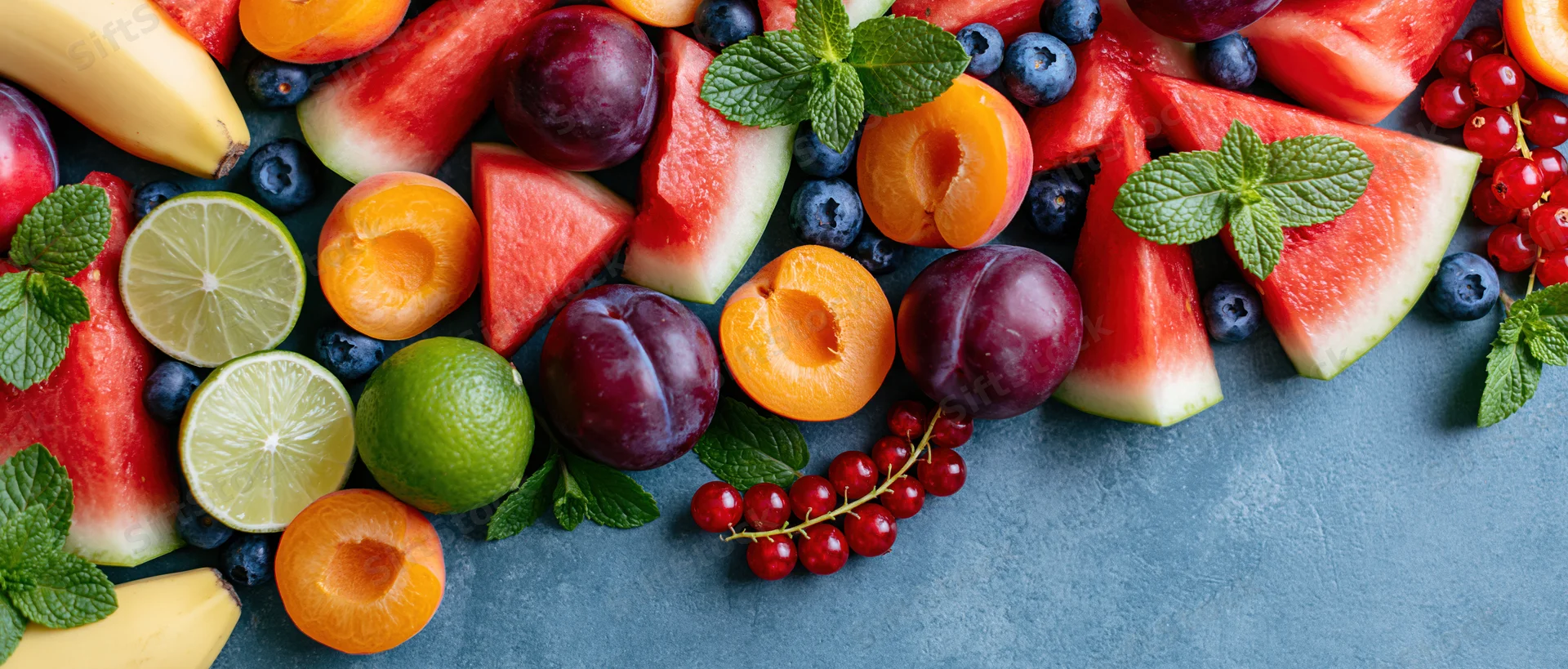Assorted sliced fruits and berries arranged on a gray slate surface with mint leaves