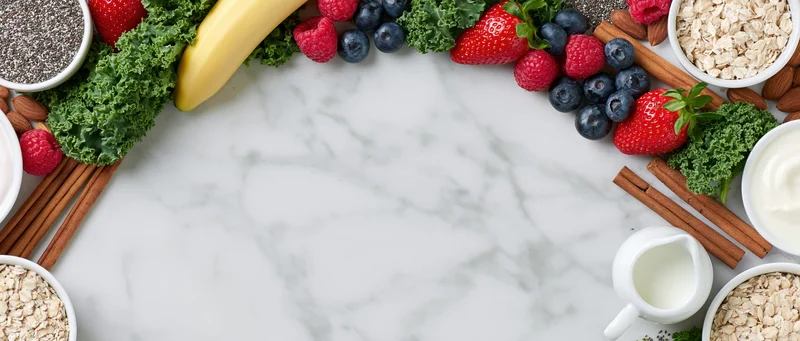 Smoothie and breakfast ingredients arranged in a border on a marble surface with copyspace in the center
