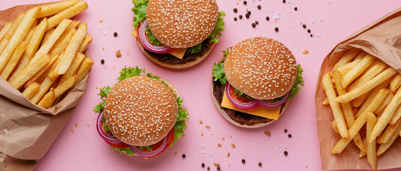 Three sesame seed burgers with lettuce tomato and onion served with french fries on pink background in paper bags