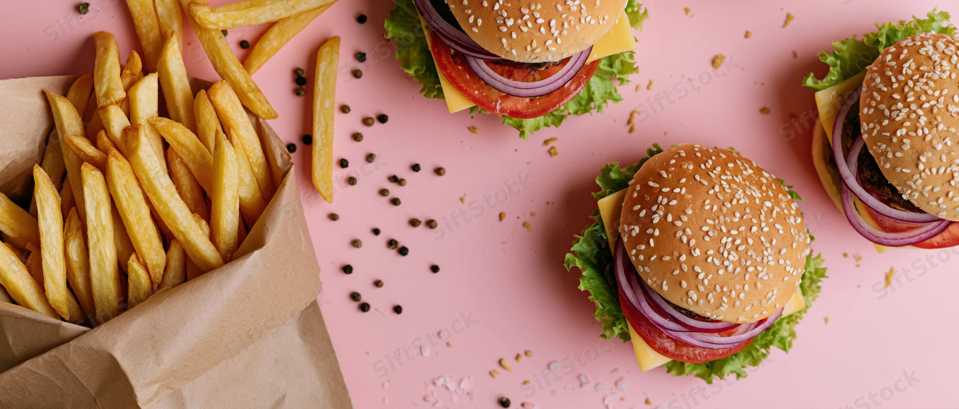 Three sesame seed burgers with lettuce tomato onion cheese and fries in a paper bag on pink surface flat lay overhead view