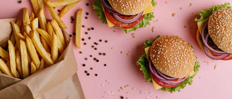 Three sesame seed burgers with lettuce tomato onion cheese and fries in a paper bag on pink surface flat lay overhead view