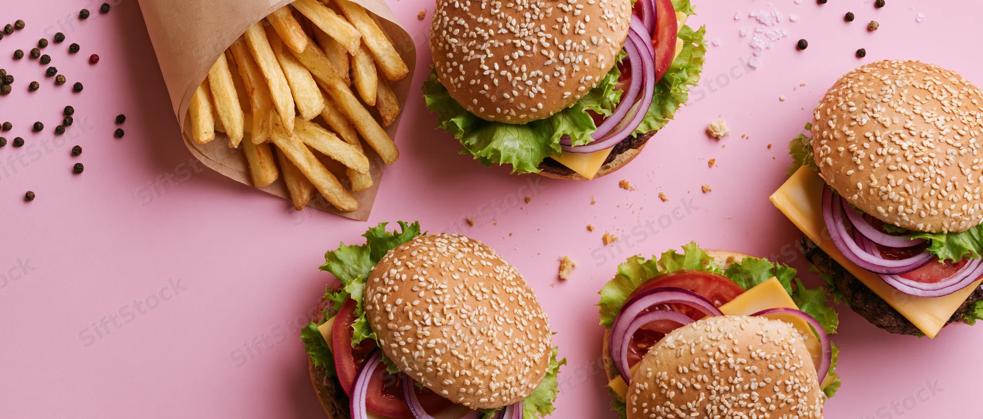 Four cheeseburgers with sesame buns and french fries arranged on pink surface with scattered seasonings and crumbs