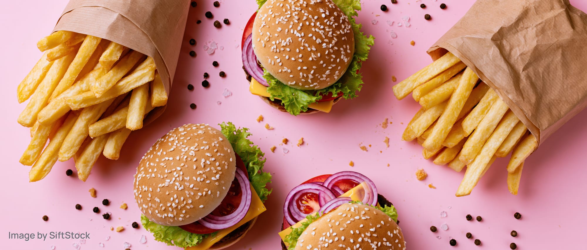 Three sesame seed burgers with lettuce tomato and cheese alongside paper-wrapped french fries arranged on a pink surface in a flat lay overhead view