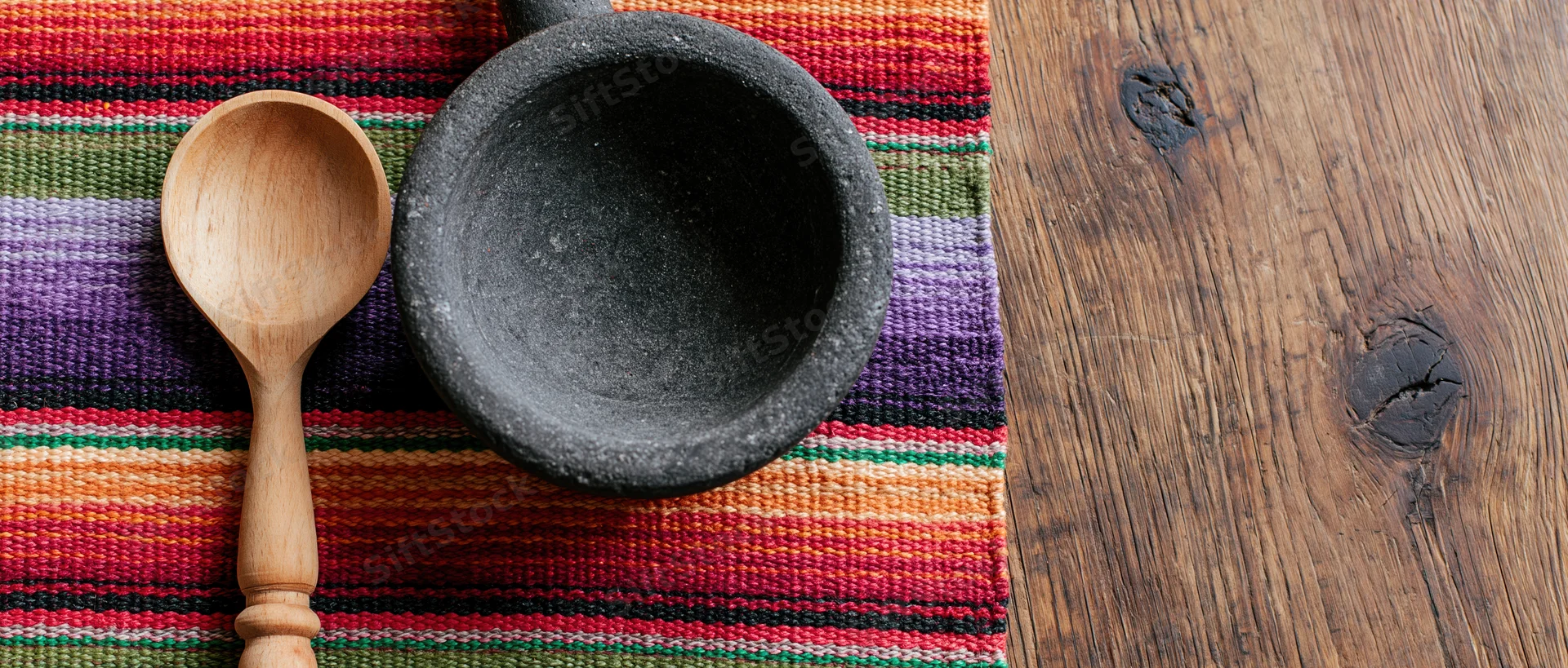 Wooden spoon and stone mortar on colorful woven textile beside wooden surface