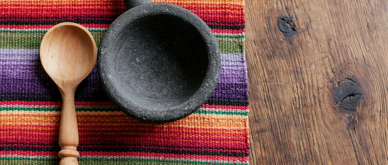 Wooden spoon and stone mortar on colorful woven textile beside wooden surface