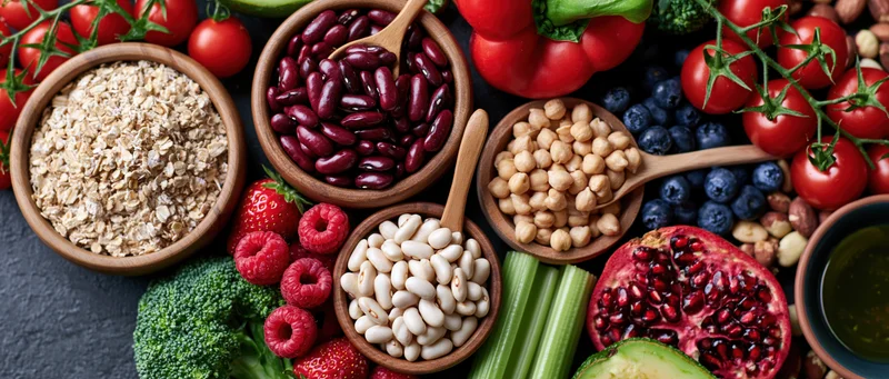 Overhead flat lay of assorted healthy foods including vegetables, fruits, beans, and grains arranged on dark surface