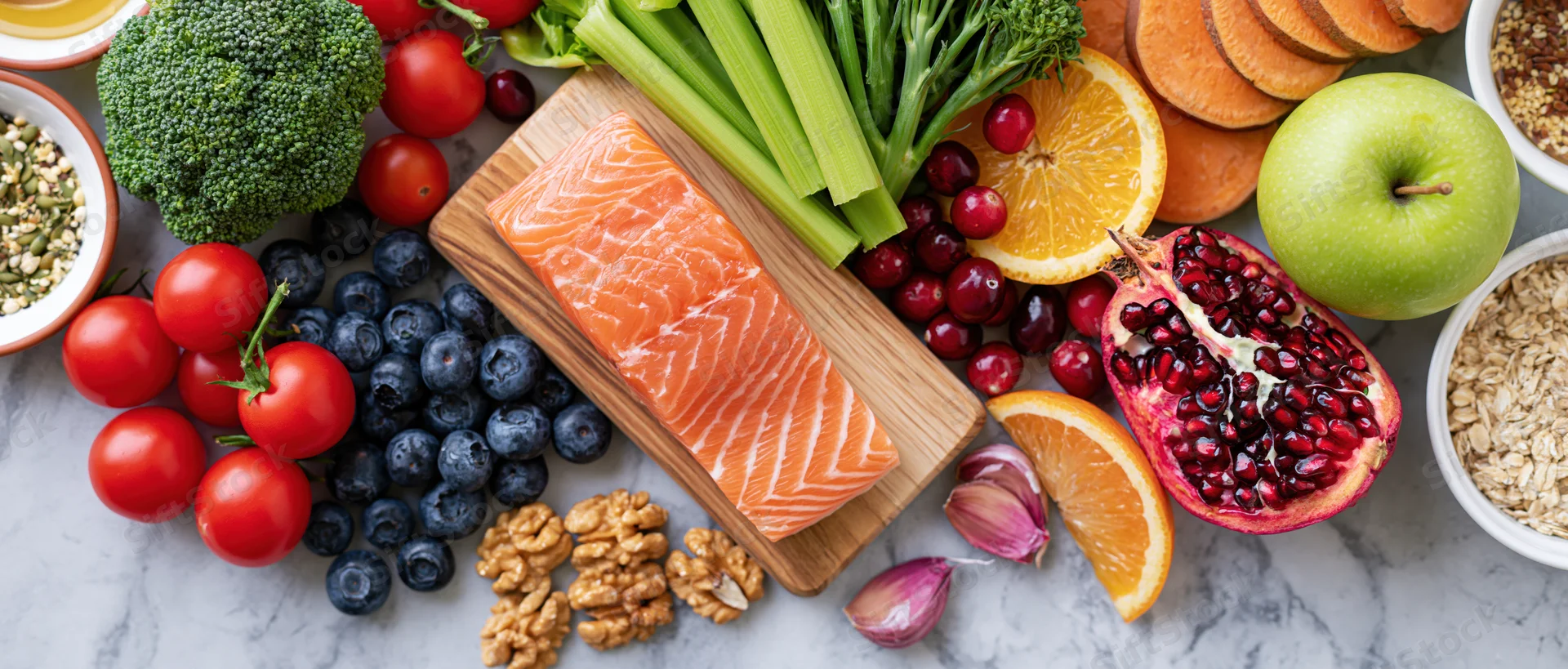 Salmon fillet on wooden board surrounded by assorted fruits and vegetables on marble surface