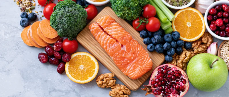 Salmon fillet on wooden board surrounded by assorted fruits vegetables nuts and seeds on gray surface