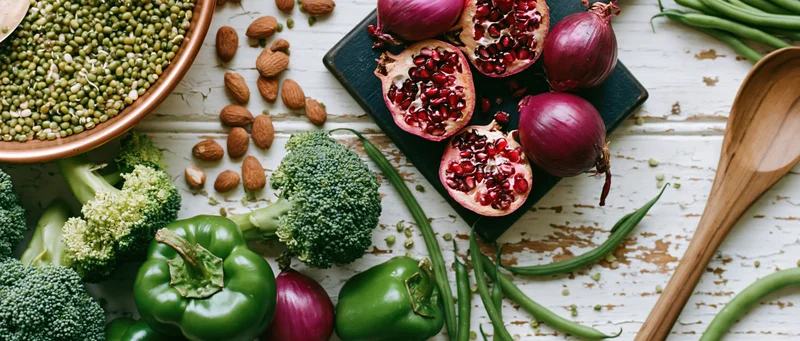 Overhead flat lay of fresh vegetables fruits and dry ingredients on a rustic white surface with a wooden spoon and cutting board
