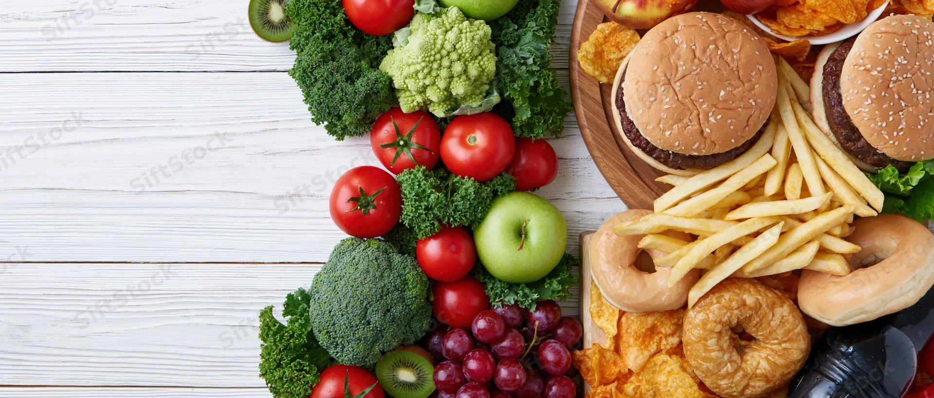 Healthy vegetables and fruits arranged next to unhealthy fast food on a white wooden table