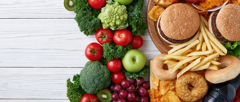 Healthy vegetables and fruits arranged next to unhealthy fast food on a white wooden table