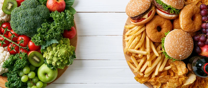Healthy vegetables and fruits on one side, junk food on the other, arranged on a white wooden table in a split composition
