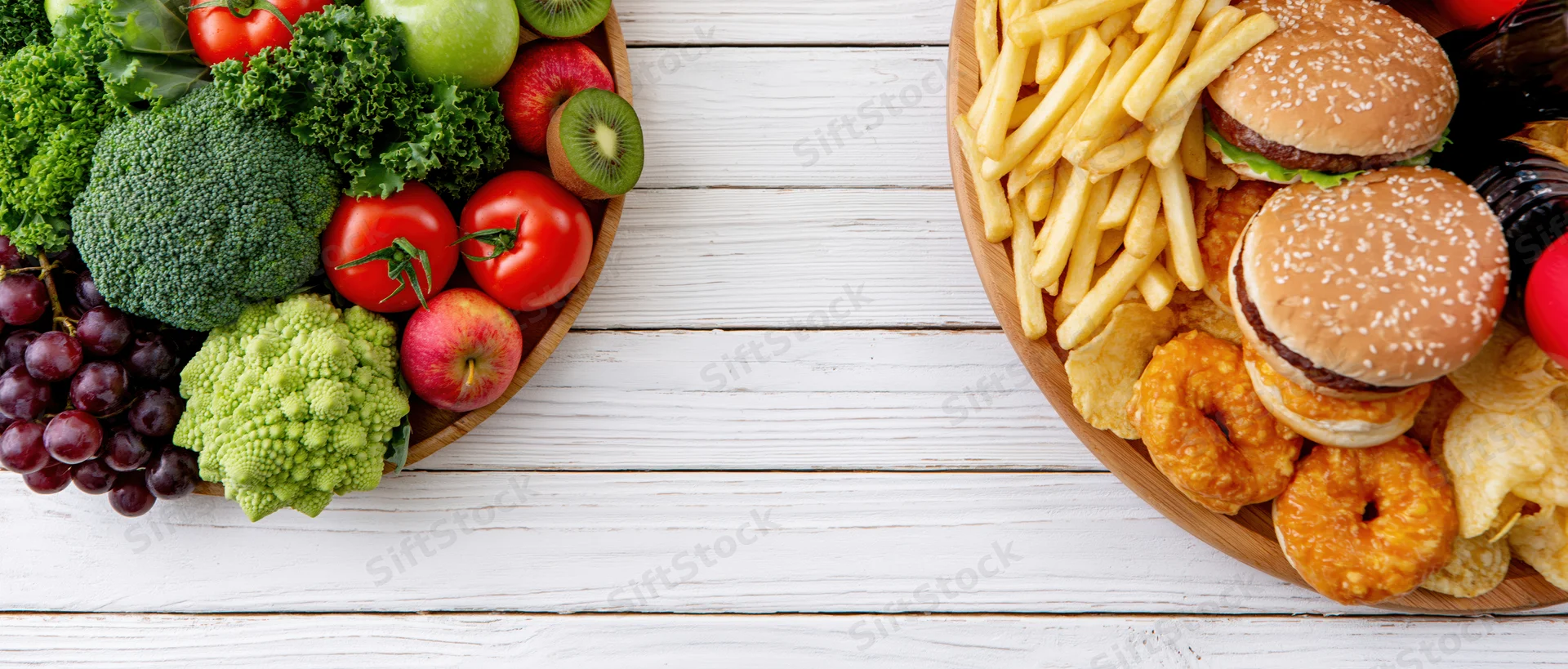 Healthy vegetables and fruits on one side, junk food with burgers and fries on the other, arranged on a white wooden table in a split composition