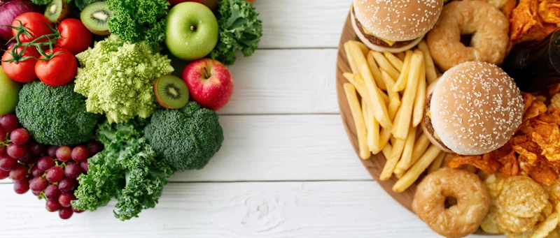 Healthy vegetables and fruits on left side of white wooden table next to junk food on right