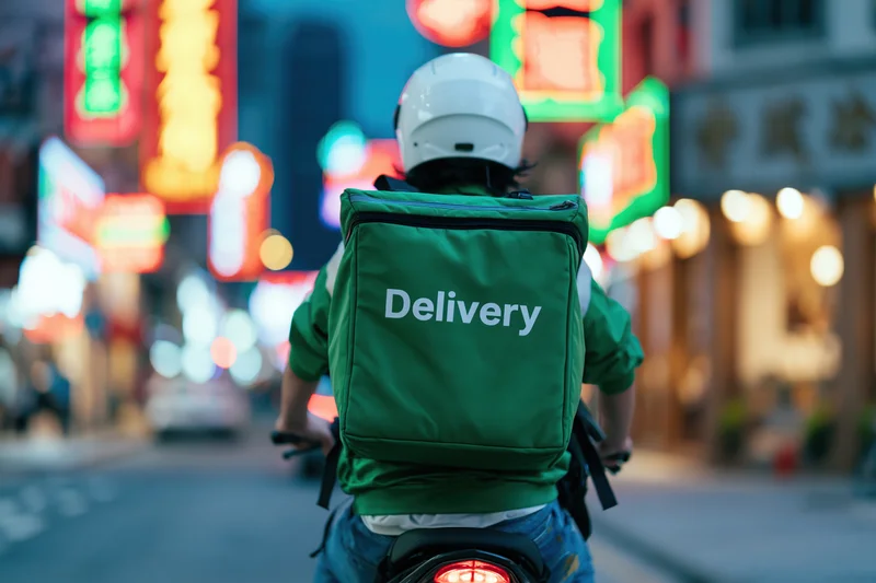 Delivery rider on motorcycle seen from behind riding through neon-lit city street at night
