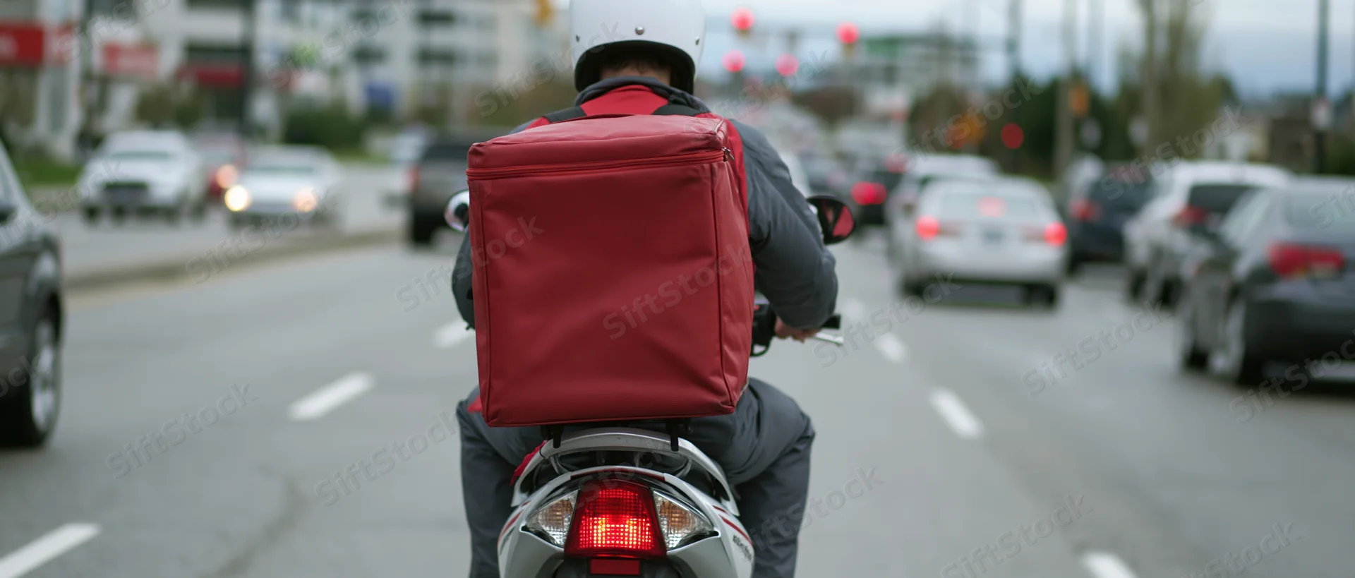 Delivery rider on scooter with red backpack riding on city street in traffic