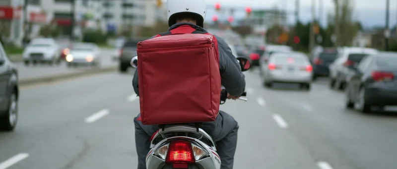 Delivery rider on scooter with red backpack riding on city street in traffic