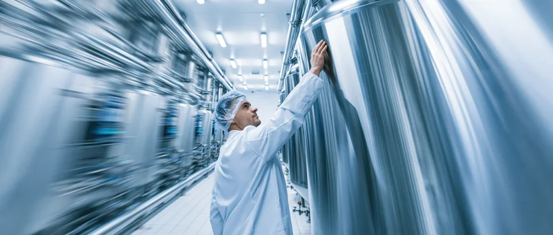 Worker in white lab coat and hairnet inspecting stainless steel tank in industrial facility