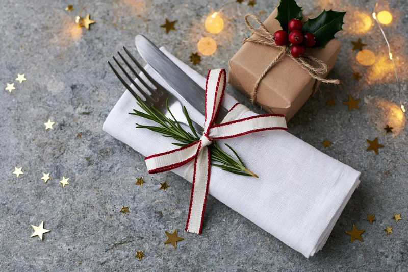 Fork and knife wrapped in a white linen napkin tied with a red and white ribbon, placed beside a small gift wrapped in brown paper with holly and berries, surrounded by golden stars and warm string lights on a gray stone surface